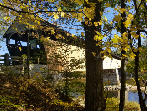 A covered bridge surrounded by autumn leaves and trees, with a clear blue sky in the background.