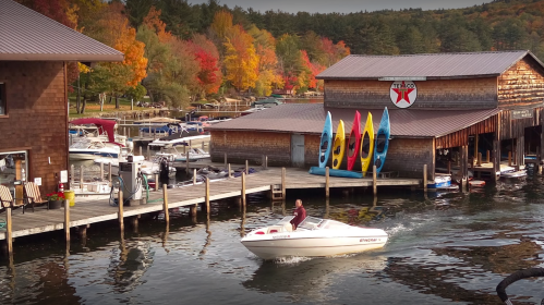 A boat speeds past a marina with colorful kayaks and autumn foliage in the background.