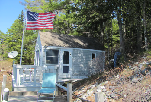 A small blue cottage with a porch, surrounded by trees, featuring an American flag and a lounge chair nearby.
