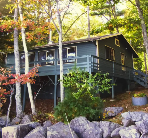 A cozy blue cabin nestled among trees, with a deck overlooking a rocky landscape and autumn foliage.