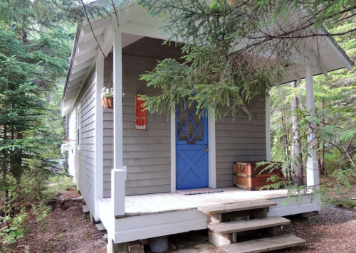 A small gray cabin with a blue door, surrounded by trees and featuring a wooden porch and steps.