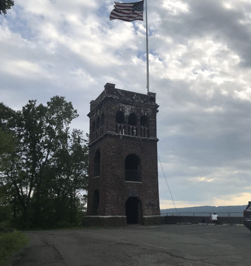 A stone tower with an American flag, surrounded by trees and a cloudy sky, near a road.