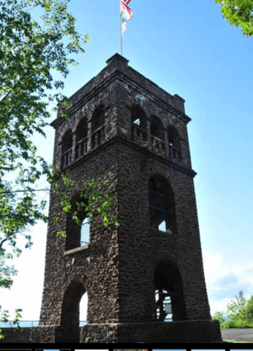 A tall stone tower with arched windows, surrounded by trees, under a clear blue sky with a flag on top.