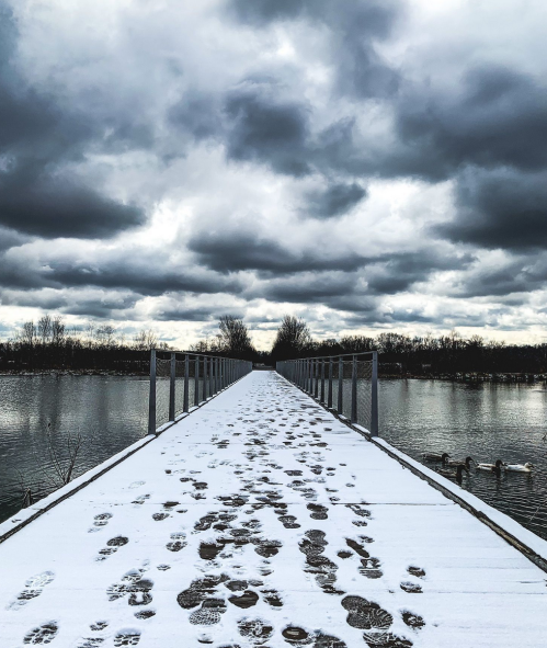 A snowy pier extends into a calm lake under a cloudy sky, with footprints leading toward the horizon.