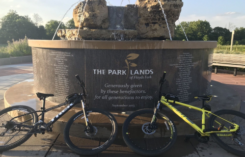 Two bicycles are parked in front of a stone fountain with a plaque about The Park Lands of Floyds Fork.