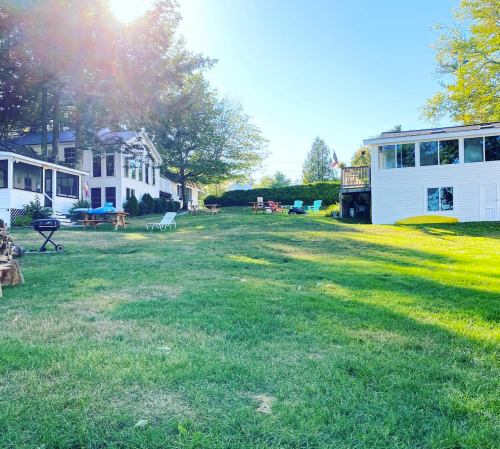 A sunny backyard with green grass, two houses, and colorful outdoor furniture scattered around.