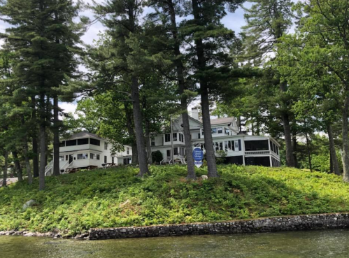 A large house surrounded by trees on a grassy hill by the water, with a clear blue sky above.