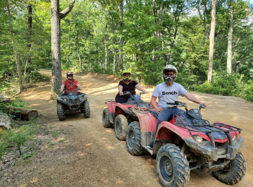 Three people riding ATVs on a dirt trail surrounded by trees, wearing helmets and protective gear.
