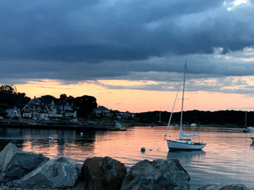 A serene waterfront scene at sunset, featuring a sailboat and charming houses along the shore, with rocky foreground.