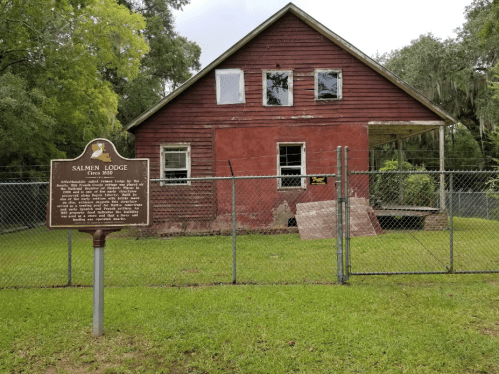 A weathered red building with a historical marker in front, surrounded by a chain-link fence and green grass.