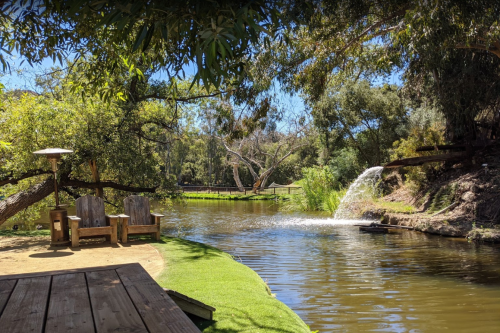A serene pond surrounded by trees, featuring two wooden chairs and a small waterfall in the background.