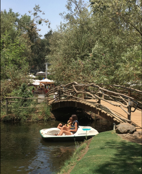 Two people relax in a paddle boat on a serene river, with a wooden bridge and lush greenery in the background.