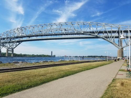 A large steel bridge spans over a river, with a pathway and grassy area in the foreground under a blue sky.