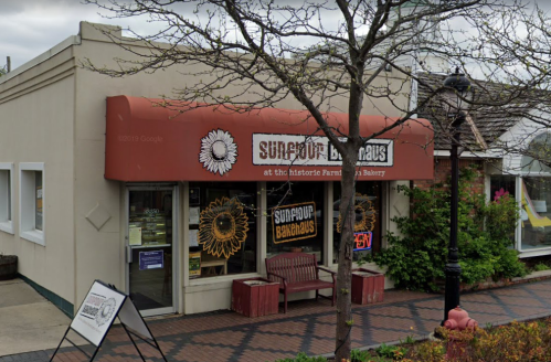 A bakery storefront with a red awning featuring sunflowers, a bench outside, and a sign indicating it's open.