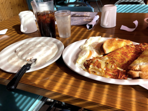 A plate with eggs, toast, hash browns, and a side of creamy gravy on a wooden table with drinks in the background.