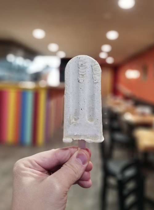 A hand holding a creamy popsicle in a colorful restaurant setting with blurred tables in the background.