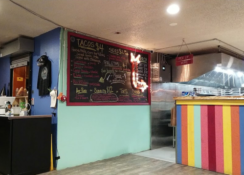 A colorful restaurant interior featuring a chalkboard menu with taco and side prices, and a kitchen area in the background.