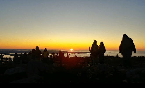 Silhouettes of people watching a sunset over the ocean, with vibrant orange and purple hues in the sky.