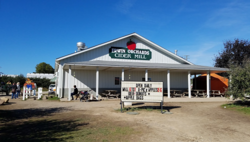 A white building with a sign reading "Erwin Orchards Cider Mill," featuring outdoor seating and a clear blue sky.
