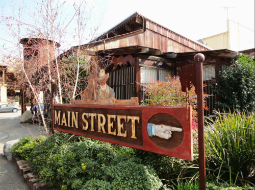 Sign for "Main Street" with a pointing hand, surrounded by greenery and rustic buildings in the background.