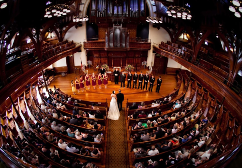 A wedding ceremony in a grand hall, with guests seated in rows and a couple at the altar surrounded by their wedding party.
