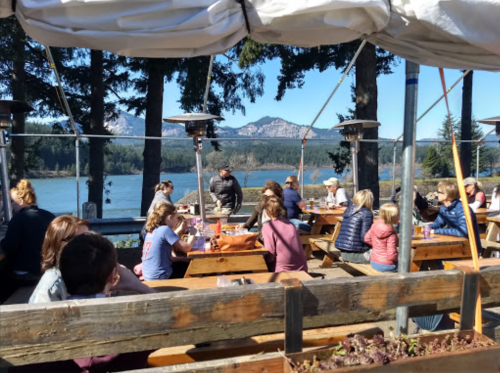 A lively outdoor dining area by a lake, with people enjoying meals and scenic mountain views in the background.