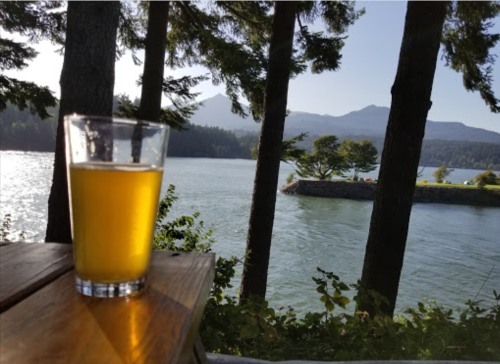 A glass of beer sits on a wooden table, with a scenic view of a river and mountains in the background.