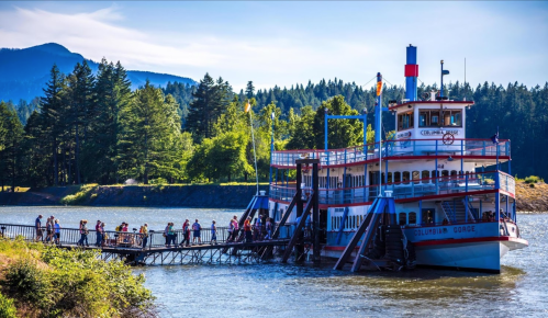 A paddleboat docked by a river with people boarding, surrounded by trees and mountains under a clear blue sky.