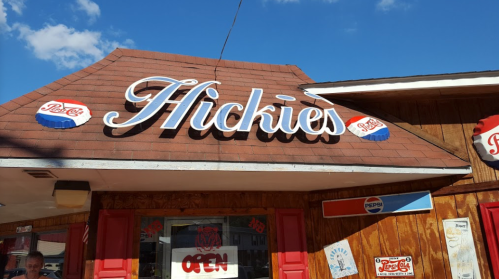 Sign for "Hickies" restaurant with a Pepsi logo, featuring a wooden exterior and bright blue sky in the background.