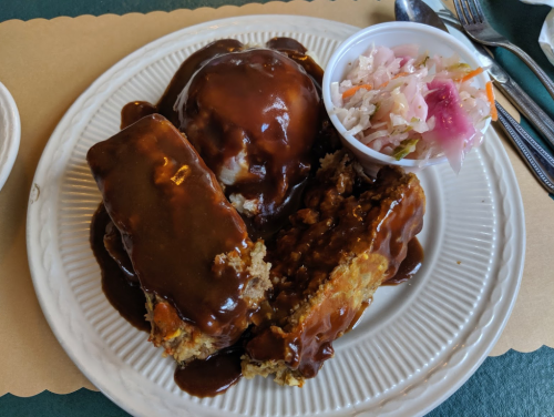 A plate of meatloaf with gravy, mashed potatoes, and a side of coleslaw in a cup.