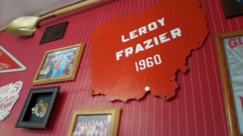 Red wall display featuring a sign that reads "Leroy Frazier 1960" alongside various framed photos and memorabilia.