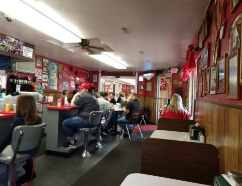 A cozy diner interior with red decor, patrons seated at booths and counters, and vintage memorabilia on the walls.