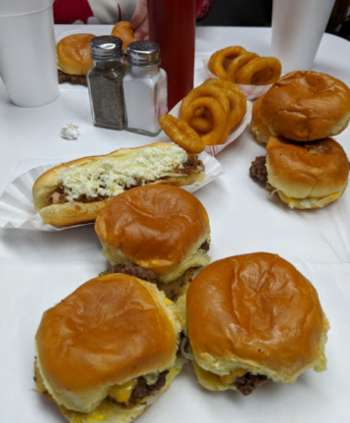 A table with burgers, a hot dog topped with coleslaw, onion rings, and condiments in the background.
