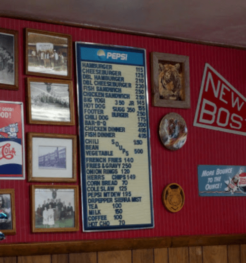 A vintage diner menu on a red wall, featuring prices for burgers, hot dogs, and drinks, alongside framed photos and decor.