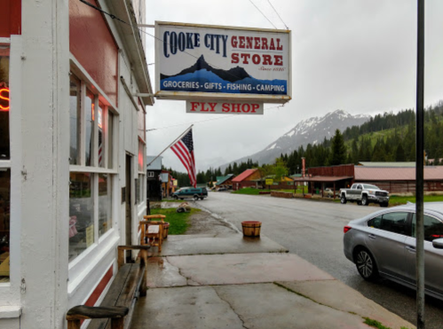 A general store sign in Cooke City, with mountains in the background and an American flag nearby on a rainy day.