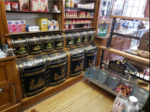 Vintage wooden counter with decorative tea canisters labeled with various types of tea, surrounded by shelves of goods.