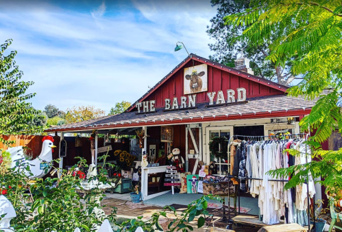 A charming red barn-style shop with a sign reading "The Barn Yard," surrounded by greenery and colorful merchandise.