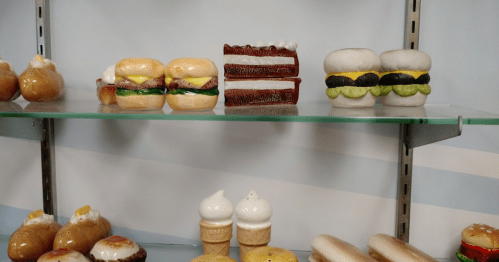 Display of colorful ceramic food items, including burgers, cake, ice cream cones, and hot dogs on glass shelves.
