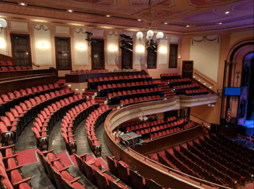 Interior of a theater featuring red seats arranged in a semi-circular layout, with ornate architectural details.