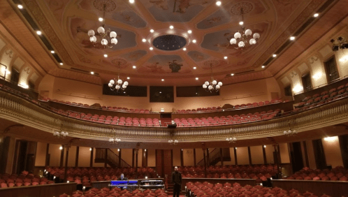 A grand theater interior with ornate ceiling, red seats, and a stage, viewed from the back.
