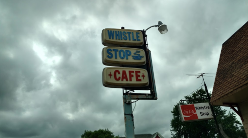 Sign for Whistle Stop Cafe under a cloudy sky, featuring retro-style lettering and a coffee cup icon.