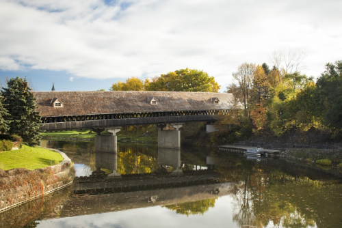 A covered bridge spans a calm river, surrounded by autumn foliage and a cloudy sky. Reflections shimmer in the water.
