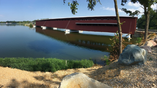 A red covered bridge spans a calm lake, surrounded by greenery and rocky shoreline under a clear blue sky.