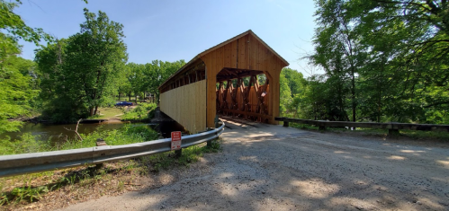 A wooden covered bridge spans a river, surrounded by lush green trees on a sunny day.