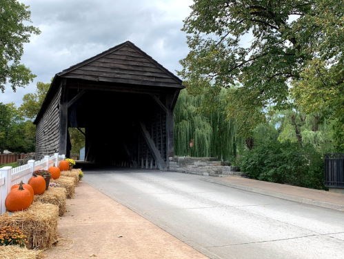 A rustic covered bridge with pumpkins and hay bales on the side, surrounded by trees and a cloudy sky.