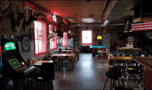 Interior of a rustic bar with wooden tables, a pool table, and vintage decor, including mounted animal heads.
