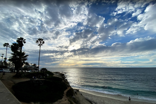 A scenic beach view with palm trees, clouds, and gentle waves under a colorful sky at sunset.