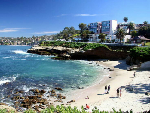 A scenic beach with rocky shores, palm trees, and a hotel overlooking the ocean, with people enjoying the sun.