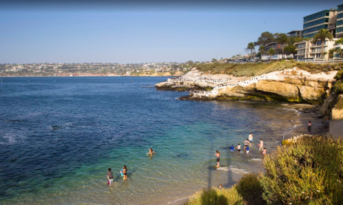 People enjoying a sunny day at the beach, with rocky cliffs and clear blue water in the background.