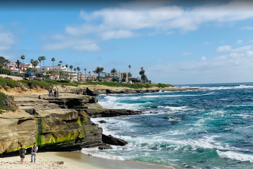 A scenic beach view with rocky cliffs, waves crashing, and people enjoying the sunny day along the coastline.